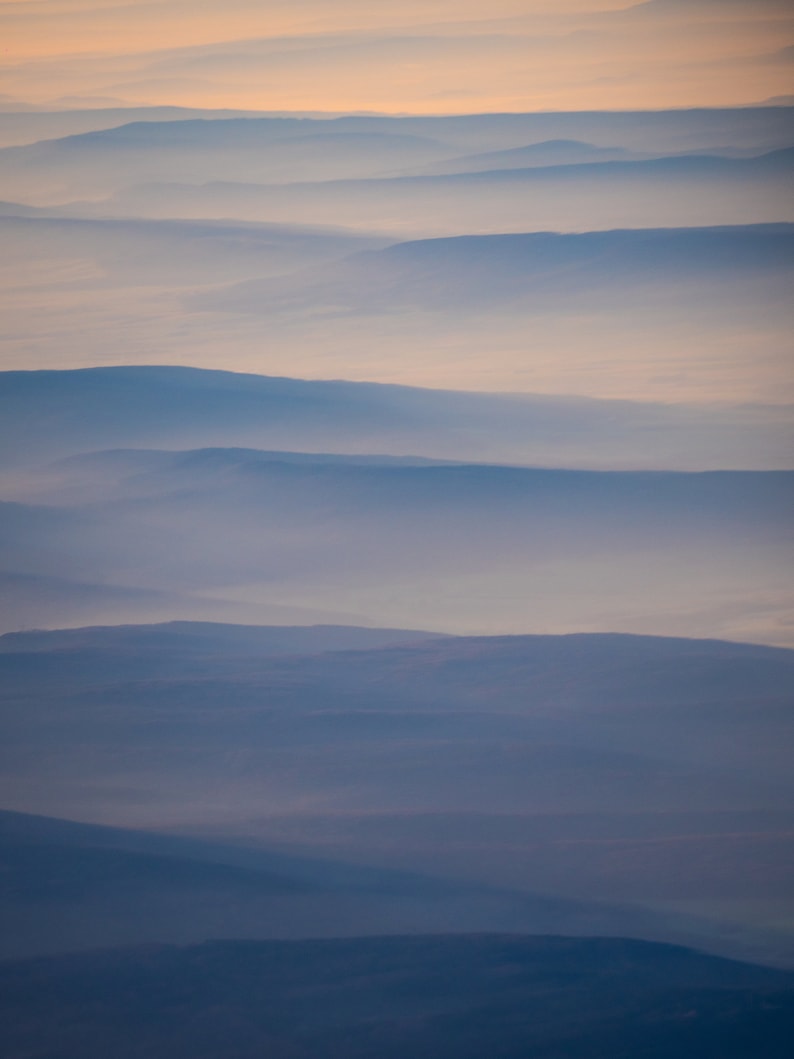 May include: An aerial view of layered mountain ranges shrouded in a soft, blue-gray mist. The sky transitions from a pale orange at the top to a deeper blue, creating a serene and atmospheric landscape.