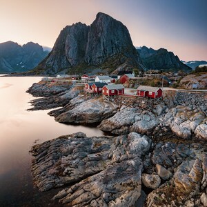 May include: A scenic view of a small red-roofed village nestled between rocky mountains and a calm body of water. The village is situated on a rocky shoreline with a few boats docked in the water.