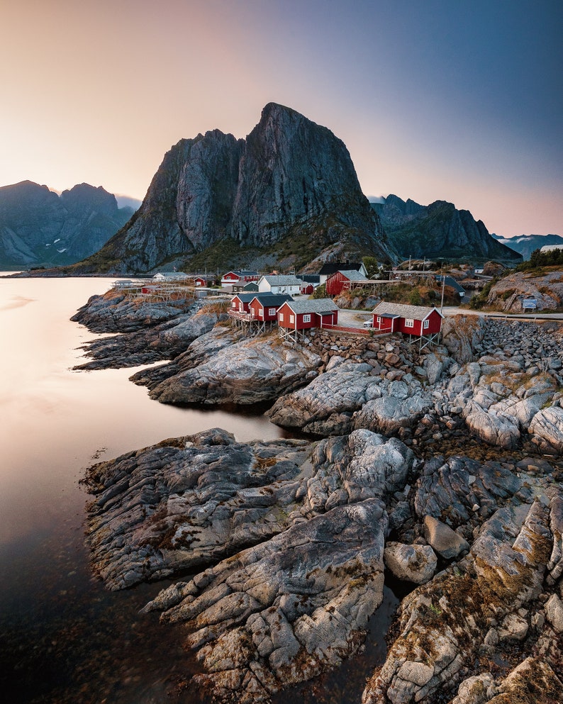 May include: A scenic view of a small red-roofed village nestled between rocky mountains and a calm body of water. The village is situated on a rocky shoreline with a few boats docked in the water.