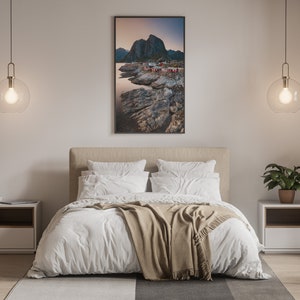 May include: A bedroom interior with a white bed, a brown blanket, and two white nightstands. There is a framed photograph of a rocky coastline with a mountain in the background hanging on the wall above the bed.
