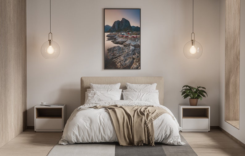 May include: A bedroom interior with a white bed, a brown blanket, and two white nightstands. There is a framed photograph of a rocky coastline with a mountain in the background hanging on the wall above the bed.