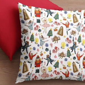May include: A white throw pillow with a colorful pattern of dancers, nutcrackers, Christmas trees, and other holiday imagery. The pillow is on top of a red pillow.