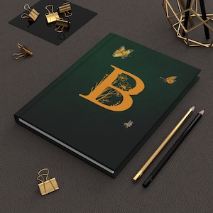 Op de afbeelding: Een donkergroen notitieboek met een gouden letter "B" ontwerp, vlinders en bloemmotieven. Gouden en zwarte potloden en gouden paperclips zijn ook zichtbaar. Het notitieboek ligt gesloten op een donkergrijze ondergrond.