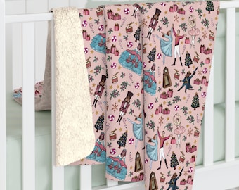 May include: A pink blanket with a repeating pattern of Christmas-themed illustrations. The design includes nutcrackers, ballerinas, Christmas trees, gifts, and clocks. The blanket has a cream-colored, plush sherpa backing.