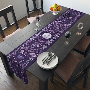 Puede incluir: Un camino de mesa morado oscuro con un diseño caprichoso que presenta lunas, mariposas y elementos florales. El camino de mesa está colocado sobre una mesa de madera oscura, creando un contraste llamativo. La mesa está puesta con platos, cubiertos y copas de vino.