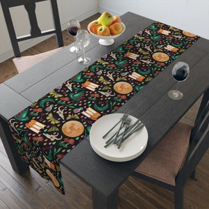 Puede incluir: Un camino de mesa negro con un patrón caprichoso de lunas, mariposas, setas y sombreros de bruja. El camino de mesa está decorado con detalles en naranja, verde, rosa y amarillo. Se encuentra sobre una mesa de madera oscura, con un frutero y copas de vino.