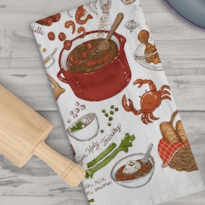 May include: A white kitchen towel with a colorful illustration of ingredients and a pot of gumbo. The text on the towel reads "Cajun Holy Trinity" and "with rice and onions".