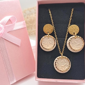 Könnte beinhalten: Ein goldfarbenes Schmuckset mit Halskette und Ohrringen, präsentiert in einer rosa Geschenkbox. Die Halskette und die Ohrringe haben ein kreisförmiges Design mit floralem Muster. Die Geschenkbox ist ebenfalls rosa und mit einem rosa Band gebunden.