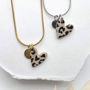 Könnte beinhalten: Zwei Halsketten mit herzförmigen Anhängern in beigem und schwarzem Leopardenmuster. Eine Kette hat eine goldfarbene Schlangenkette und einen kleinen runden Goldanhänger, die andere eine silberfarbene Schlangenkette und einen kleinen runden Silberanhänger.