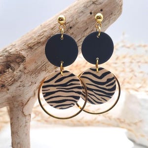 Könnte beinhalten: Ohrringe zum Hängen mit einer schwarzen Scheibe, einer beigefarbenen Scheibe mit Zebraprint und einem goldenen Reifen. Die Ohrringe sind an einem goldenen Stecker befestigt. Die Ohrringe werden auf einem Stück Treibholz präsentiert.