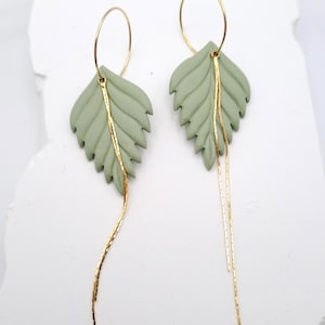 Könnte beinhalten: Ein Paar ohrringe in Form von salbeigrünen Blättern mit goldfarbenen Akzenten. Die Ohrringe haben ein strukturiertes Blattdesign mit einer goldenen Kette, die von unten herabhängt. Vor weißem Hintergrund.