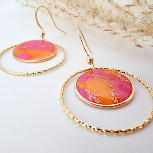 Könnte beinhalten: Goldfarbene Creolen-Ohrringe mit strukturiertem Design. Jeder Ohrring hat einen kreisförmigen Anhänger mit einem rosa-orangenen Marmormuster, akzentuiert mit Goldflocken. Die Ohrringe haben einen Hakenverschluss.