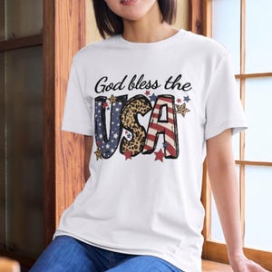 Könnte beinhalten: Weißes T-Shirt mit dem Text "God bless the USA". Das Wort "USA" ist mit einer Kombination aus Mustern gestaltet: blauen Sternen, Leopardenmuster und rot-weißen Streifen. Das Shirt hat einen Rundhalsausschnitt und kurze Ärmel.