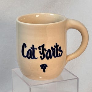 以下が含まれることがあります： ベージュ色のセラミックマグカップ。曲がったハンドルが付いています。「Cat Farts」の文字が濃い青色の筆記体で書かれており、テキストの下には小さな青色のグラフィックがあります。マグカップは透明な正方形のスタンドに展示されています。