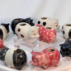 Pode incluir: Uma coleção de vasos de cerâmica em forma de porco em várias cores e padrões. Os vasos são em tons de preto, branco e vermelho, alguns com bolinhas ou efeitos marmorizados. Cada vaso tem uma abertura circular no topo.