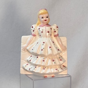 May include: A vintage ceramic figurine of a girl in a white dress with pink floral accents. The figurine has blonde hair and is mounted on a rectangular base. The dress features a tiered skirt and puffed sleeves.