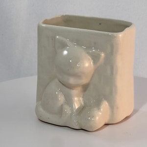 May include: White ceramic planter shaped like a cat sitting with its paws crossed. The planter has a rectangular opening at the top for planting.