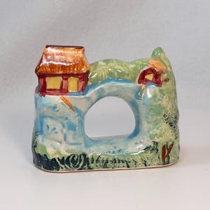 May include: A ceramic napkin ring featuring a whimsical village scene. The ring has a bridge-like design with a house on each side, painted in shades of red, yellow, green, and blue. The ring is a decorative item.