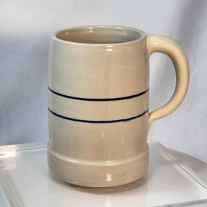 May include: A beige ceramic mug with a handle and two dark blue horizontal stripes. The mug has a cylindrical shape with a slightly flared base and a rounded handle. The mug is likely used for drinking beverages.