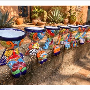 May include: A row of six ceramic planters, each shaped like a pair of legs with feet dangling over a ledge. The planters feature vibrant, multi-colored hand-painted patterns in shades of blue, orange, red, yellow, and green with white interiors.