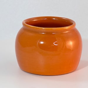May include: A small, round, orange ceramic pot with a glossy finish. The pot has a wide opening and a slightly curved body. It is a simple, yet elegant piece, suitable for various uses.