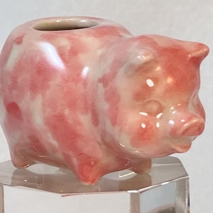 May include: A ceramic pig ornament in pink and white hues. The pig has a small opening on its back, possibly for a plant or decoration. The figurine is displayed on a clear, geometric stand. The measurements are in centimetres.