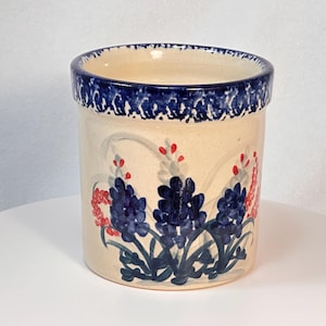 May include: A cylindrical ceramic utensil holder with a cream-colored base and a dark blue rim. The holder is decorated with blue and red floral designs. The rim has a dark blue, textured pattern.