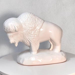 May include: A ceramic figurine of a bison in a light pink color. The bison is standing on a textured base. The figurine has detailed fur and facial features. The sculpture is a decorative item.
