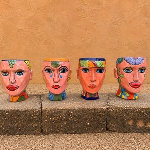 May include: Four ceramic head-shaped planters arranged in a row. Each planter features a unique hand-painted face with vibrant colors, including pink skin tones, blue eyes, and red lips, decorated with various floral and geometric patterns.