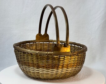 May include: A woven metal basket with a rounded shape and two arched handles. The basket is made of a gold-toned metal, with a woven pattern on the sides and a solid metal handle attached with brass-colored brackets.