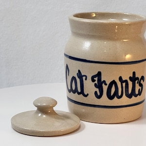 May include: A beige ceramic jar with a lid, featuring the words "Cat Farts" in dark blue script. The jar has a dark blue band around the top and bottom. The lid is off to the side.