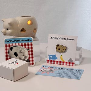 May include: A collection of pig-themed items, including a ceramic pig night light, a pin, a card with a pig illustration, and a small white box with "thank you" printed on it. The pin features a polka-dotted pig design.