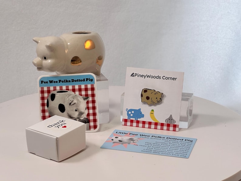 May include: A collection of pig-themed items, including a ceramic pig night light, a pin, a card with a pig illustration, and a small white box with "thank you" printed on it. The pin features a polka-dotted pig design.
