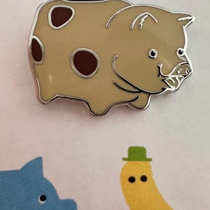 May include: A tan-colored piggy bank pin with brown spots and a silver outline. The pin has a smiling face and a slot on top for coins. The image also shows a blue pig and a yellow banana with a green hat.