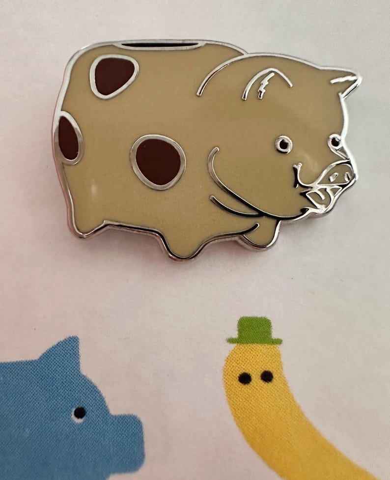 May include: A tan-colored piggy bank pin with brown spots and a silver outline. The pin has a smiling face and a slot on top for coins. The image also shows a blue pig and a yellow banana with a green hat.