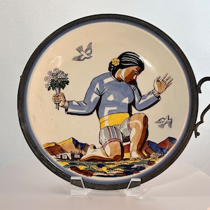 May include: A ceramic plate with a hand-painted design of a person kneeling and holding a bouquet of flowers. The plate has a blue and white border and a pewter rim. The plate has a handle on the side.