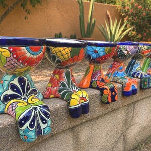 May include: A row of vibrantly painted ceramic planters with a unique, footed design. Each planter features a colorful floral and geometric pattern in shades of blue, red, orange, yellow, and green. The planters are displayed on a stone wall.