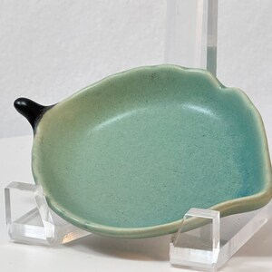 May include: A small, light green ceramic dish with a black handle. The dish is shaped like a leaf and has a slightly rounded bottom.