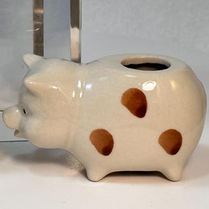 May include: A ceramic pig figurine with brown spots. The pig has a small opening on its back. The figurine is off-white with brown spots and black eyes. The pig is a decorative item.