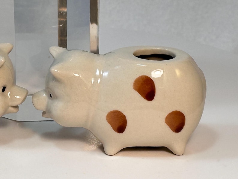 May include: A ceramic pig figurine with brown spots. The pig has a small opening on its back. The figurine is off-white with brown spots and black eyes. The pig is a decorative item.