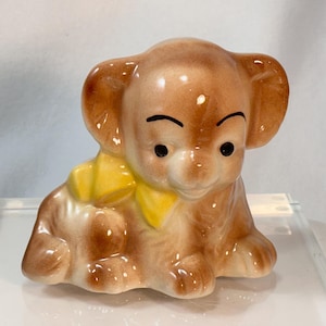May include: A ceramic puppy figurine in shades of brown and tan, with a yellow bow. The puppy has black eyes and a sweet expression. This decorative item is a charming addition to any collection.