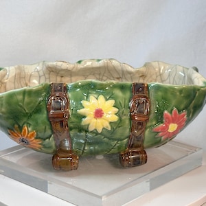 May include: A ceramic bowl with a leaf-like shape, painted green with brown accents. The bowl features a floral design with yellow and red flowers. Brown straps are on the front of the bowl.