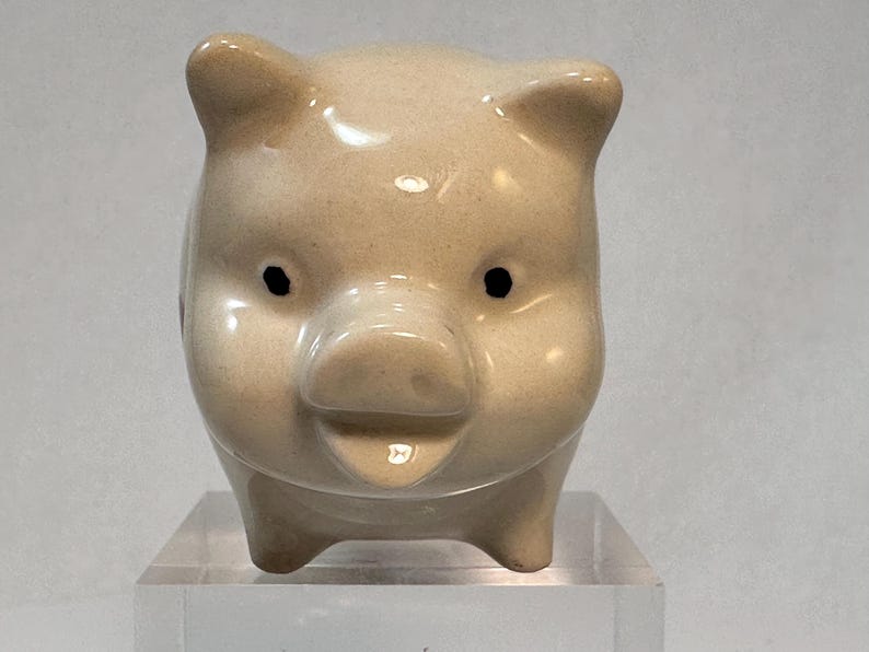May include: A small, ceramic piggy bank in a light beige color. The pig has a rounded body, small ears, and black eyes. It is standing on a clear, rectangular base. The piggy bank is a classic item for saving money.
