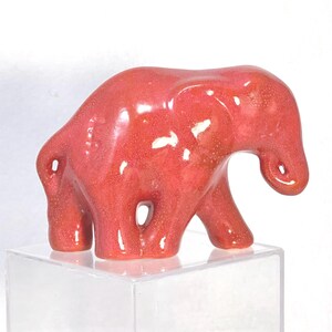 May include: A small, red ceramic elephant figurine with a glossy finish. The elephant has a trunk, ears, and four legs. The figurine is displayed on a clear, square stand. The elephant is a decorative item.