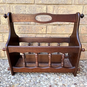 May include: A dark brown wooden magazine rack with a decorative oval cutout in the back. The rack has a slatted front and rounded edges.