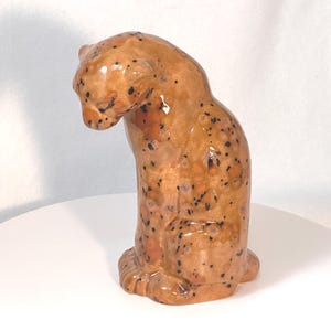 May include: A ceramic figurine of a seated leopard with a glossy finish. The sculpture features a tan base color with dark brown and black spots distributed across its body. The leopard is depicted in a resting position with its head bowed downward.