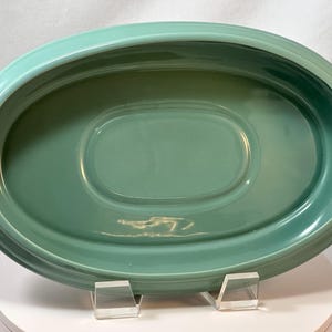 May include: A vintage, oval-shaped serving platter in a soft, seafoam green color. The ceramic dish has a slightly raised rim and a recessed oval center. The platter is displayed on a white surface with clear acrylic supports.
