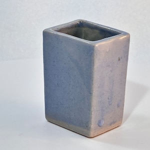 May include: A rectangular, light blue ceramic vase with a square opening. The vase has a slightly textured surface and a pale pink base. The interior is a darker shade of blue. The vase is likely used for holding flowers or as a decorative item.