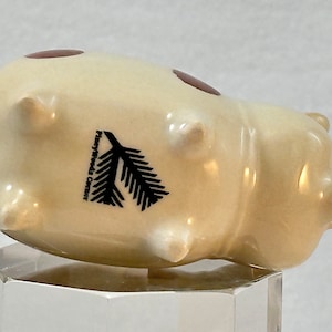 May include: A ceramic pig figurine in a cream color, with two brown oval accents and four small protrusions. A black fir tree graphic and the text "Pineywoods Center" are printed on the side. The figurine is displayed on a clear, geometric stand.