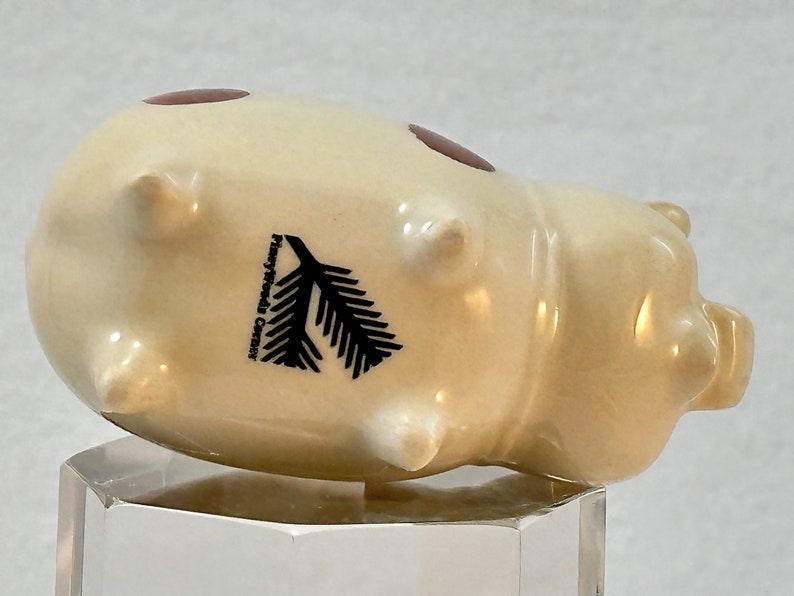 May include: A ceramic pig figurine in a cream color, with two brown oval accents and four small protrusions. A black fir tree graphic and the text "Pineywoods Center" are printed on the side. The figurine is displayed on a clear, geometric stand.